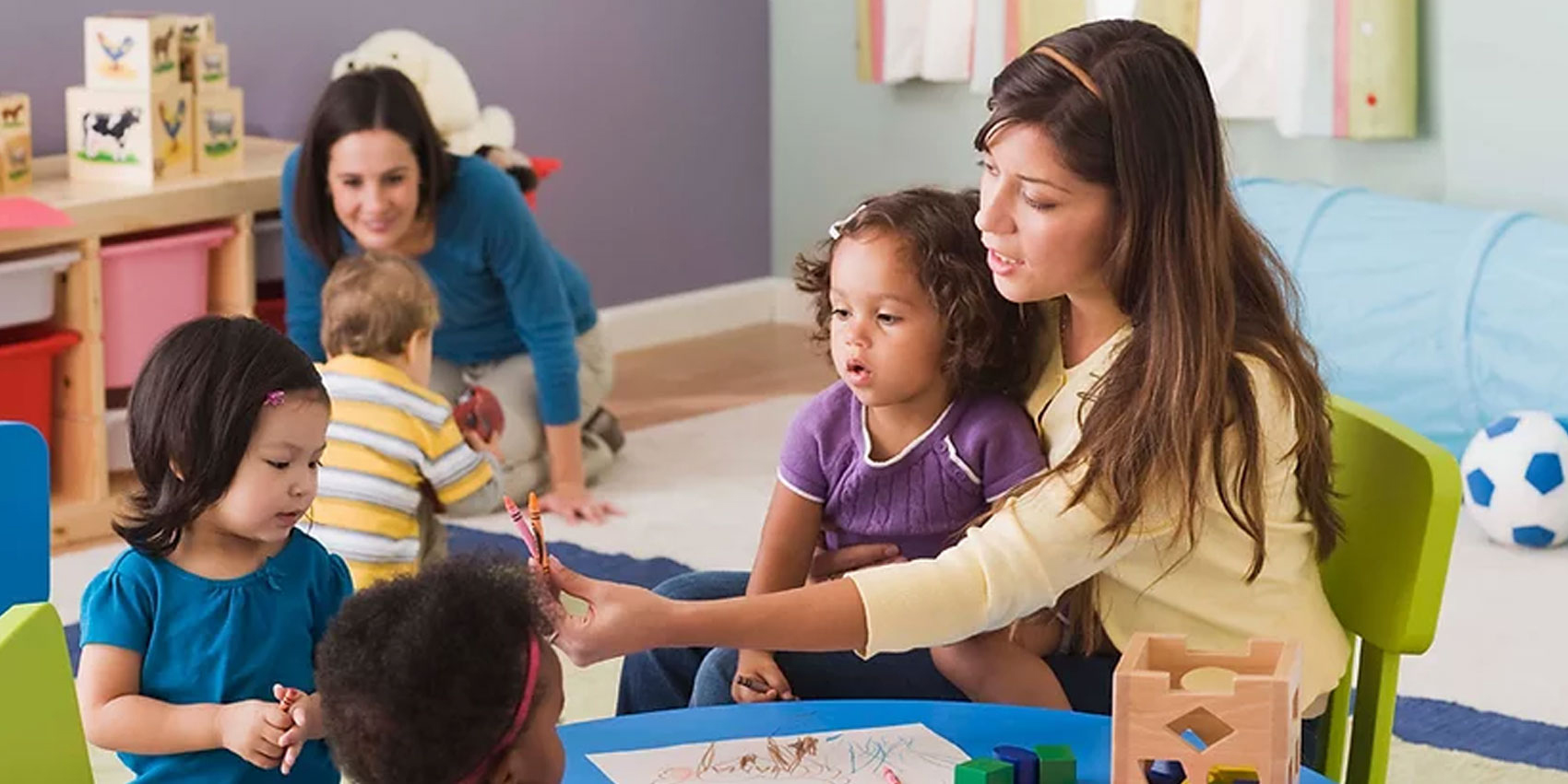 "Adult engages with several young children during a classroom activity at a low table. One child sits on the adult’s lap while the adult holds up a crayon or writing tool for the children to observe. Additional children and an adult are visible in the background, along with classroom furniture and learning materials, highlighting a collaborative early childhood learning environment."