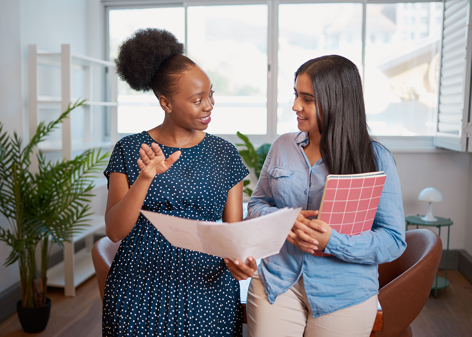 "Two individuals review a document together in an office setting. One person gestures toward the document while the other holds a notebook. Large windows, indoor plants, and office furnishings are visible in the background, highlighting collaboration, coaching, and professional support services."